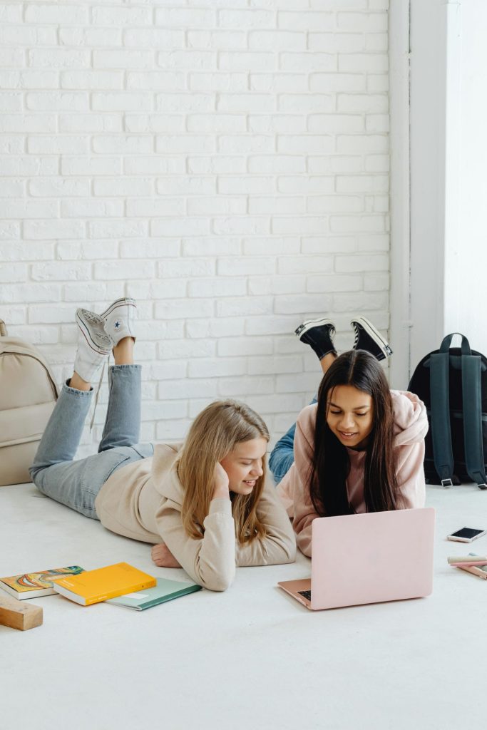 Two teenage girls studying together indoors, lying on the floor with a laptop and books.